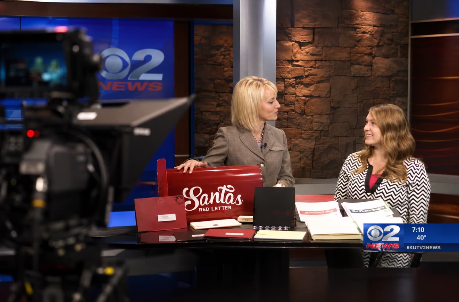 Crystal Stapley being interviewed on KUTV CBS2 News with a Santa's Red Letter mailbox display in front of her on the news desk. Camera in the foreground, news anchor on the left.